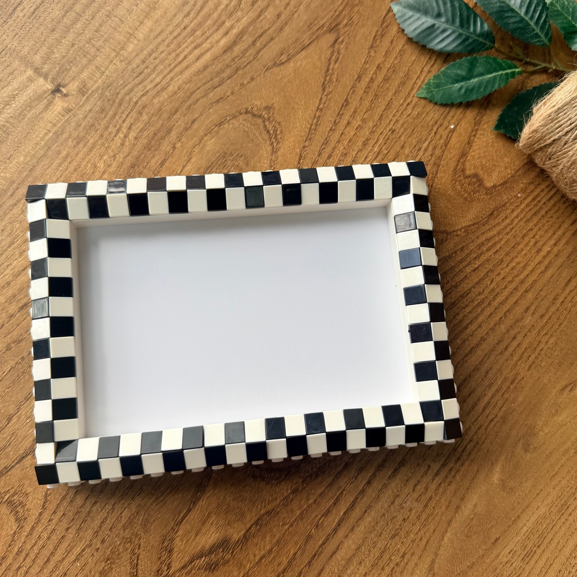 A rectangular picture frame with a black and white checkered border, displayed on a wooden tabletop next to a roll of twine and green leaves.