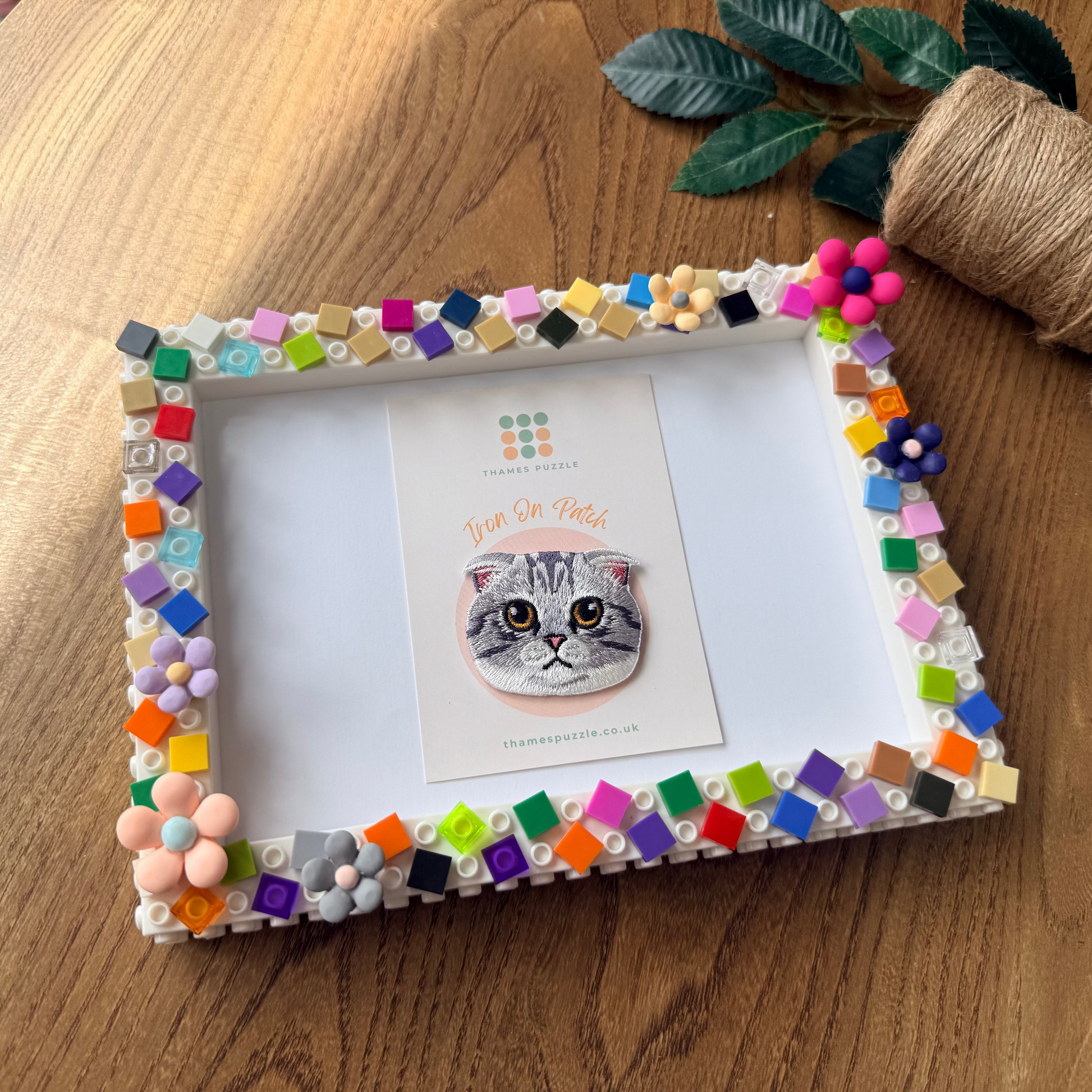 A handmade rectangular picture frame decorated with colorful mosaic tiles and beads, holding a card with a cat illustration, styled on a wooden tabletop with a plant and twine nearby.