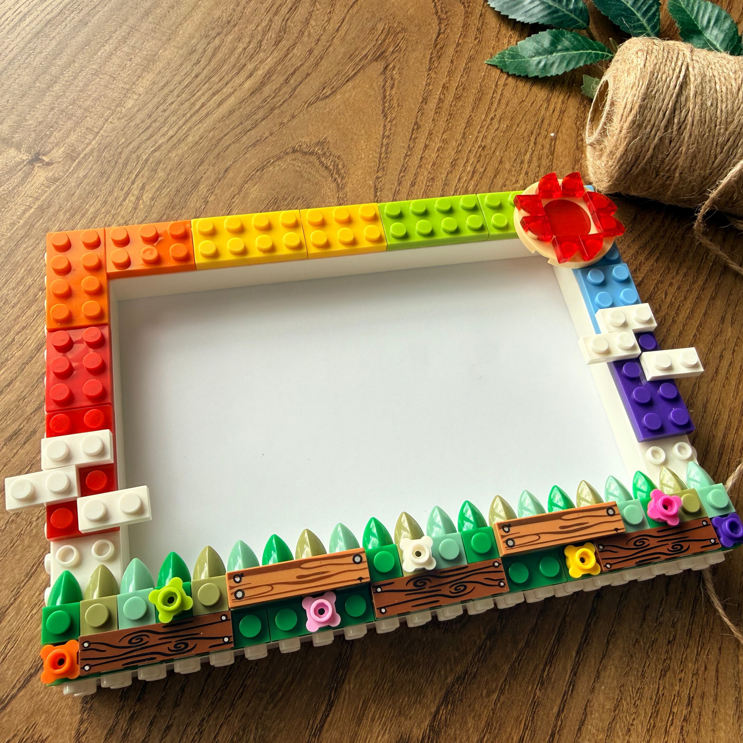 A handmade picture frame crafted from colorful LEGO bricks, featuring a rainbow gradient on one side and green leaves, flowers, and wood textures on the other. The frame is styled on a wooden tabletop next to a spool of twine and small green leaves.
