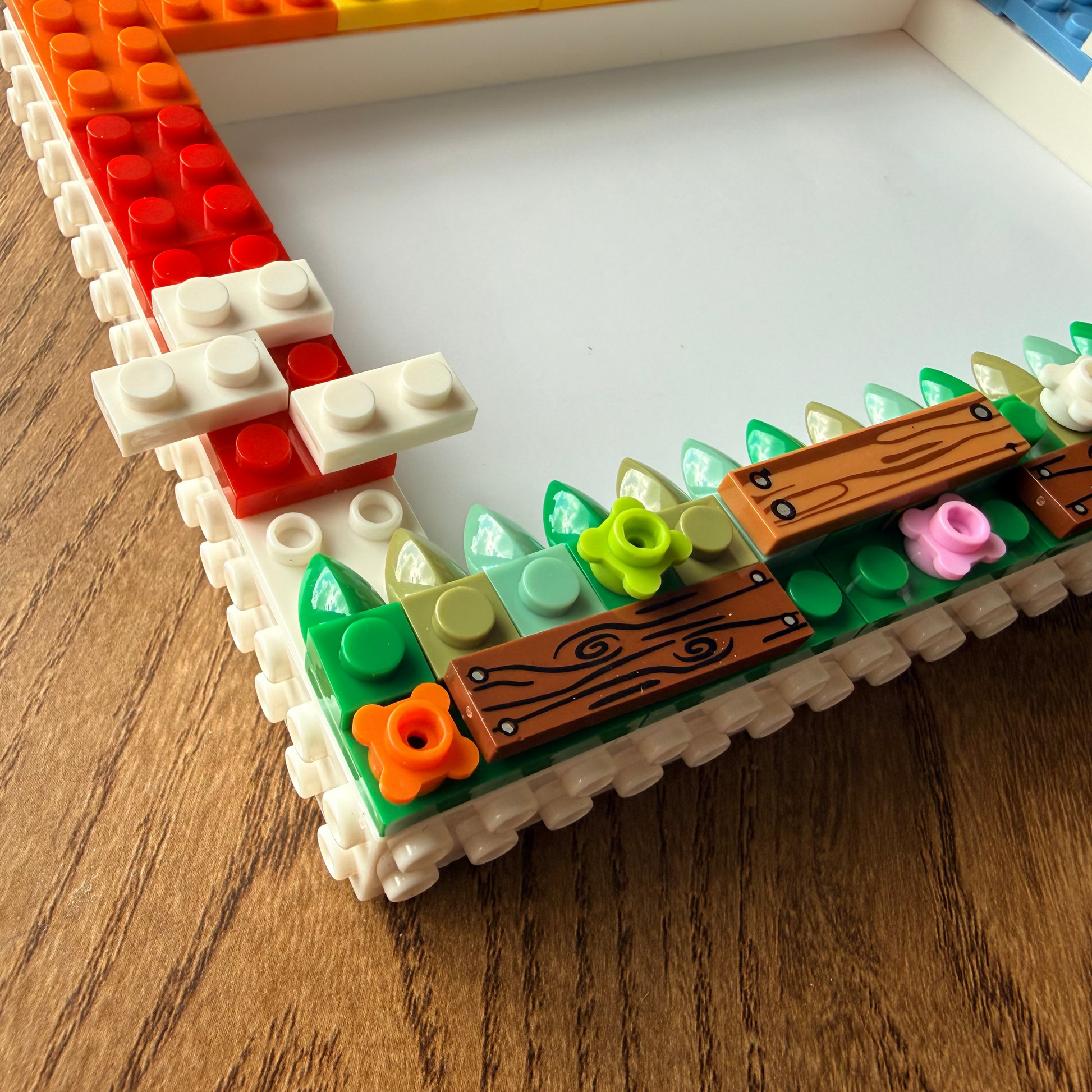 A close-up of a custom LEGO-style picture frame decorated with colorful bricks, green leaf pieces, brown wood-grain accents, and small flower pieces, shown on a wooden tabletop.
