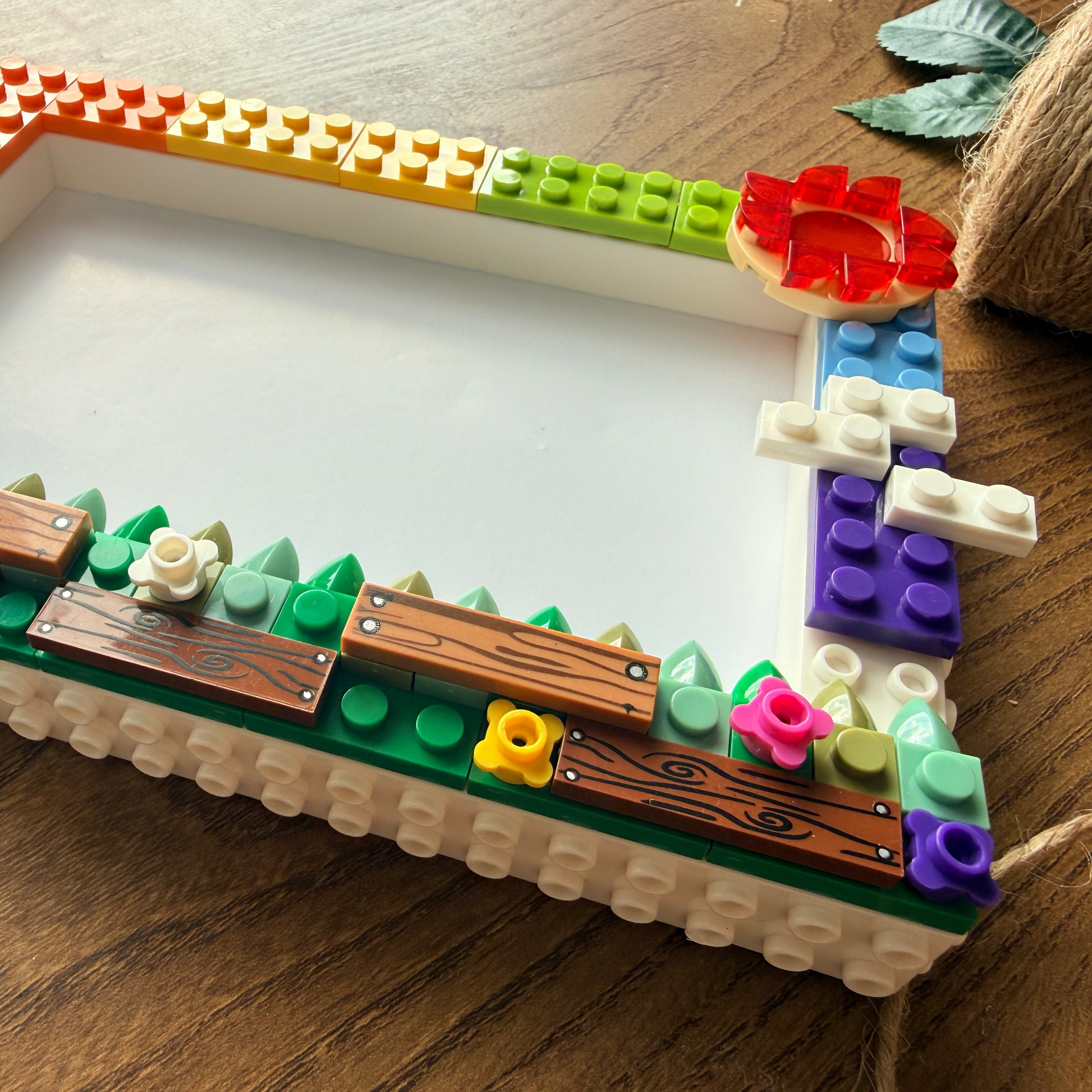 A rectangular picture frame crafted from LEGO-style bricks, decorated with a rainbow border, green base, wooden planks, small flowers, and a red flower on the right side, placed on a wooden tabletop.