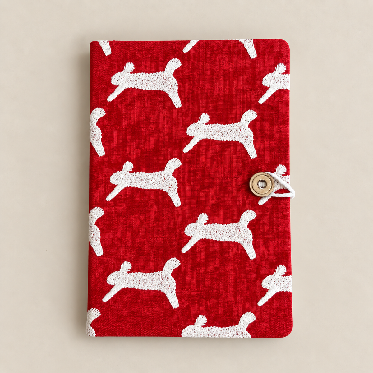 A red fabric notebook or journal cover with a repeating white dog silhouette pattern and a small metal snap closure on the right, photographed against a plain light beige background.